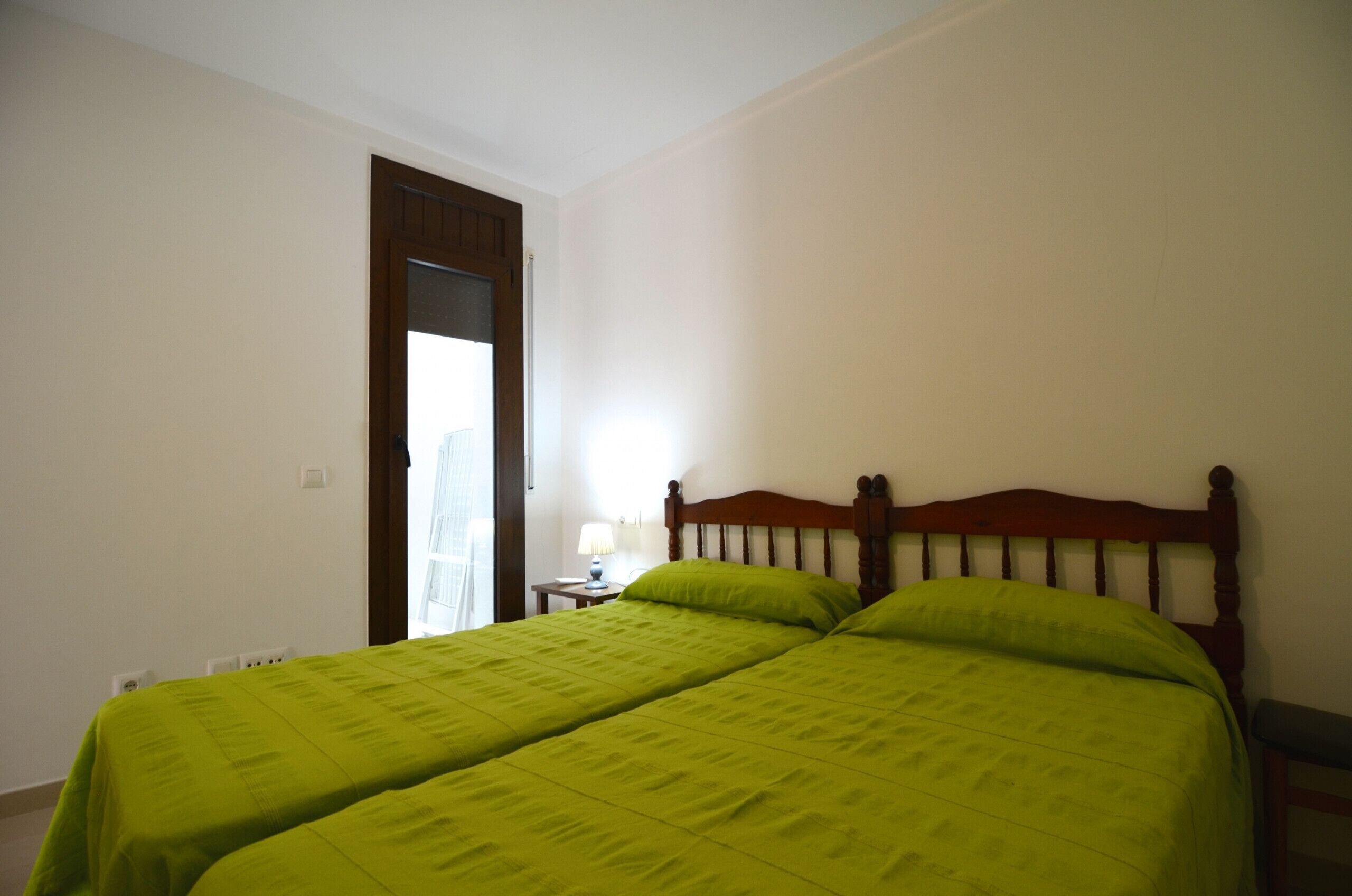 A room with two beds, a wooden headboard, a nightstand with a lamp, and a door leading to another room.