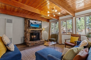 Un salon chaleureux avec une cheminée en pierre, des poutres apparentes en bois au plafond et de grandes fenêtres donnant sur l’extérieur.