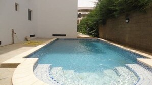 En rektangulär swimmingpool med en kant av kakel, omgiven av en låg mur och en vit byggnad.