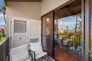 Un balcón con puerta de cristal, dos sillas de mimbre con cojines y vista a una zona residencial con palmeras.