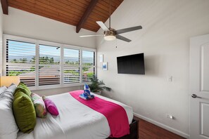 Un dormitorio con una cama, ventilador de techo, televisión y vistas al exterior a través de amplias ventanas.