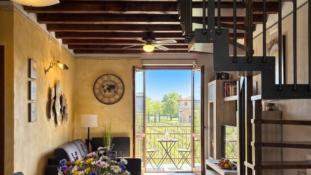 A dining area with a wooden table, chairs, a vase of flowers, wine glasses, and a bottle of wine. There is a clock on the wall, a ceiling fan, and a view of the outdoors through a glass door. - SG Family - Girasole Apartment (Peschiera del Garda)