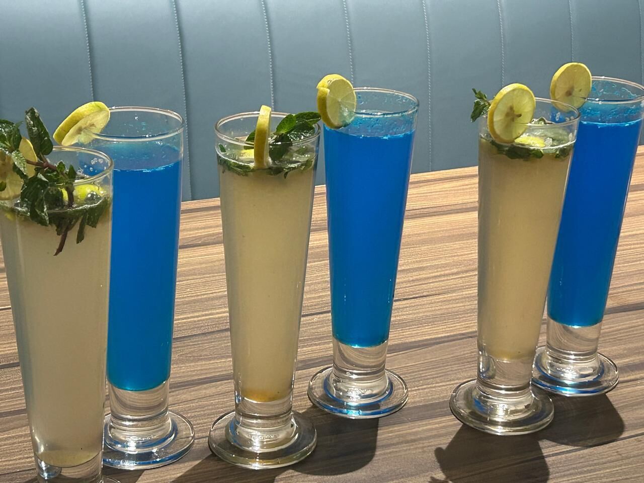 Cuatro vasos altos con bebidas de color azul, adornados con rodajas de limón y hojas de menta, sobre una mesa de madera.
