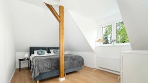A bedroom with a sloped ceiling, a bed with a grey headboard, a wooden post, a white radiator, and a window with a view of trees.