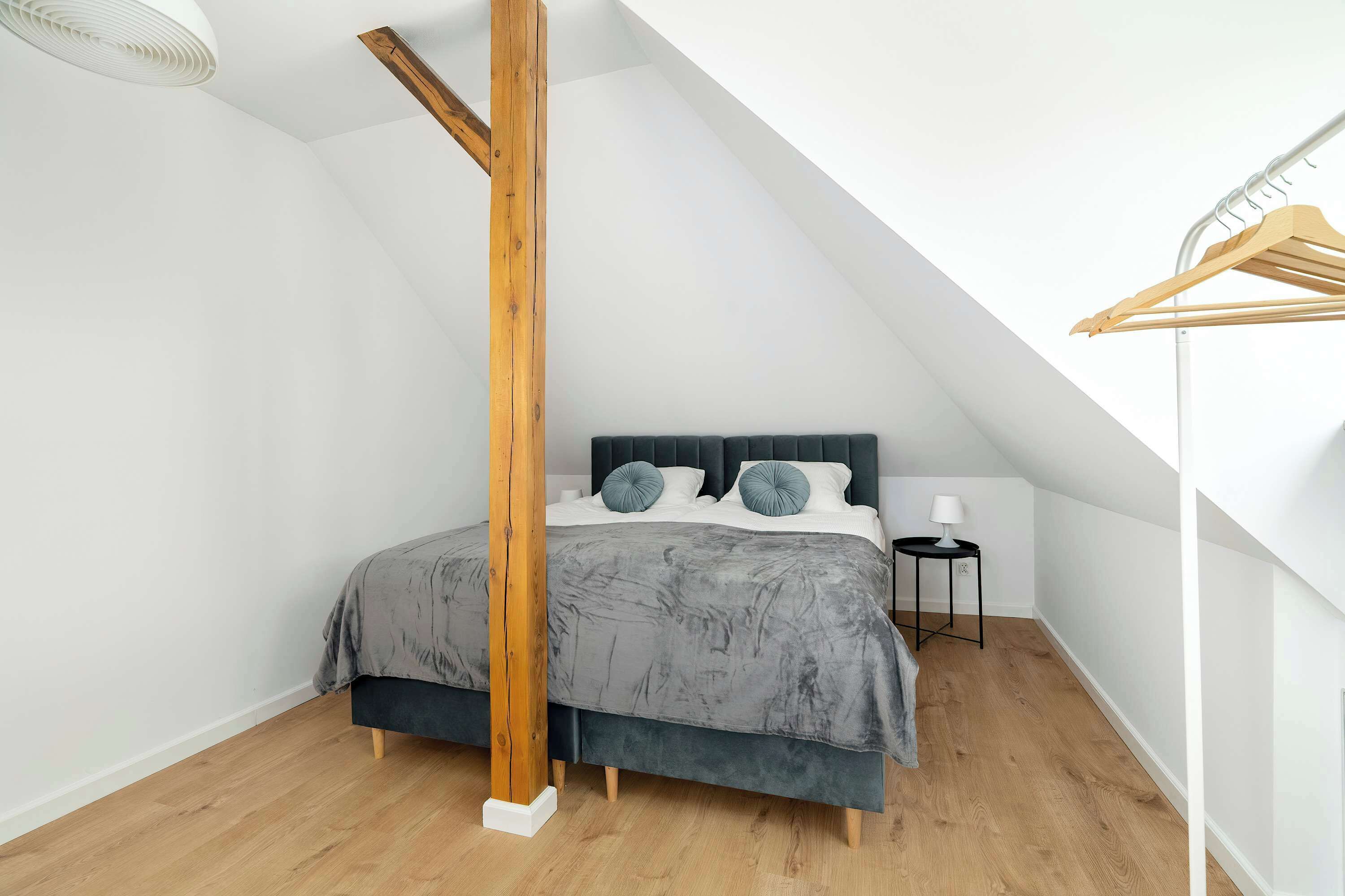 Ein Schlafzimmer mit schrägverdachter Decke, einem Bett mit dunklem Kopfteil, einem Holzmast, einem kleinen Nachttisch mit Lampe und Holzboden.