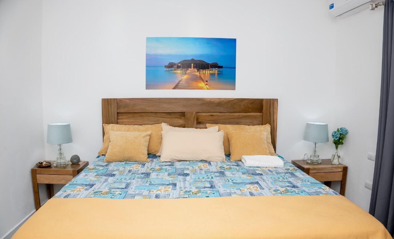 Een slaapkamer met een houten hoofdbord, een bed met een beddekleed met een patroon, twee nachtkastjes met lampen, en een schilderij van een pier.