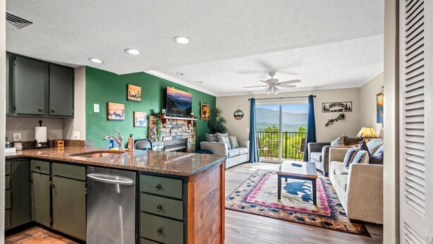A modern kitchen with green cabinets, a stainless steel dishwasher, and a large island. There is a dining area with a sofa and a coffee table, and a balcony with a view of the mountains. - Mountain Views Indoor Pool & Sauna Fireplace (Gatlinburg)