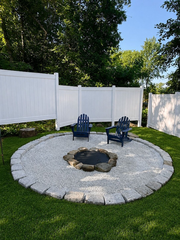 A circular stone fire pit surrounded by a white fence, with two Adirondack chairs facing the fire pit.