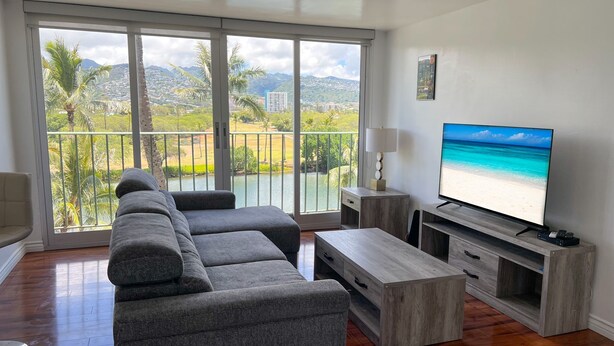 Un salón espacioso con un televisor de pantalla plana grande, un sofá modular gris, una mesa de centro de madera y un balcón con vistas a una playa y montañas.