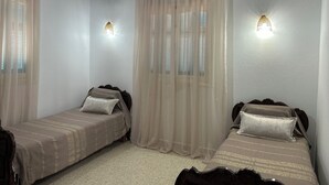 Une chambre avec deux lits simples, des rideaux légers, des lampes fixées au mur et un ventilateur de plafond.
