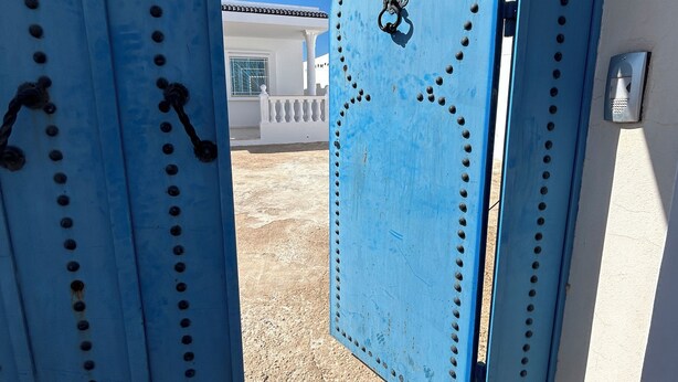 Une porte métallique bleue ornée d’un emblème circulaire, qui donne accès à un bâtiment aux murs blancs et doté d’un balcon.