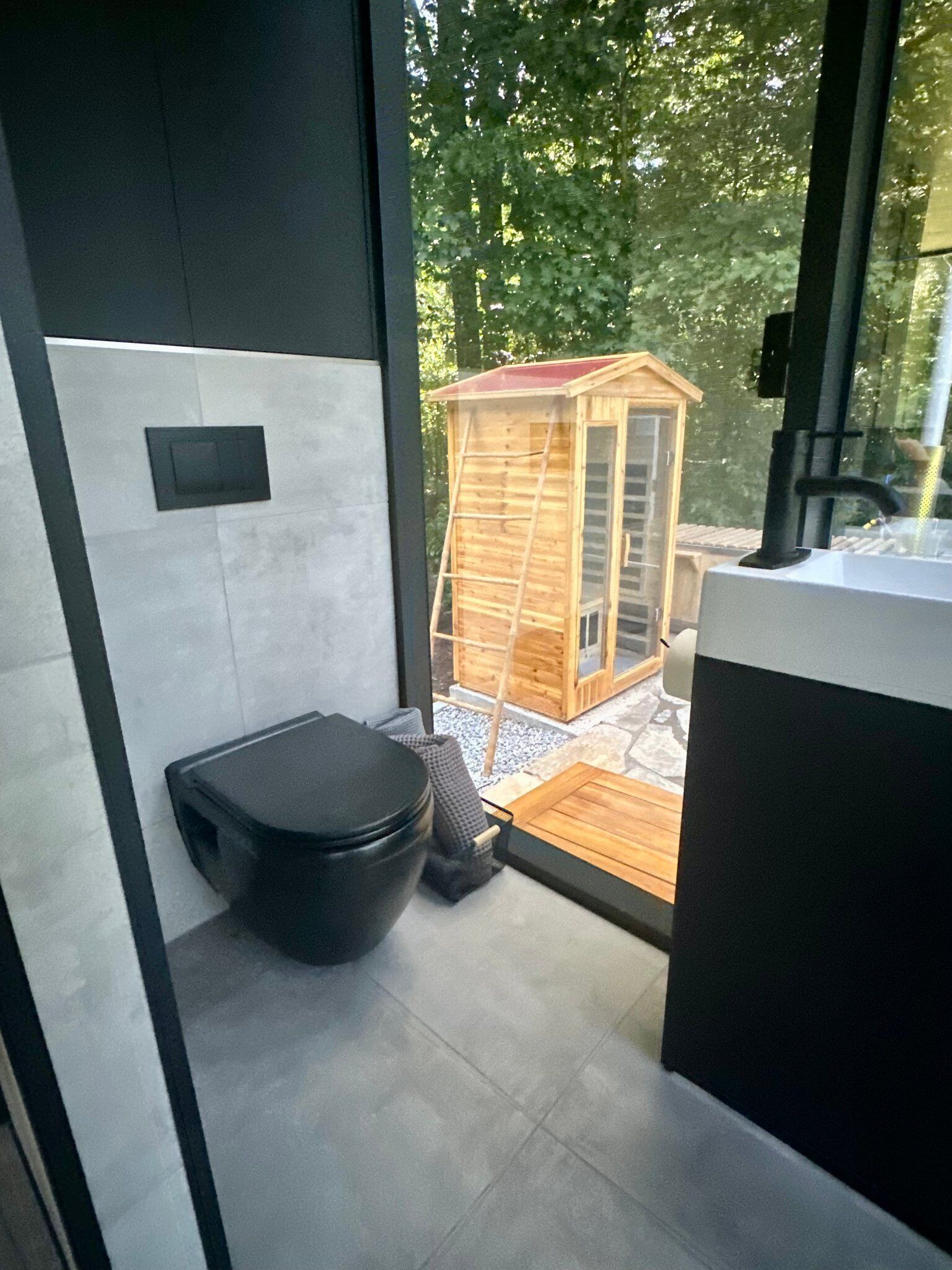 A modern bathroom with a toilet, a wooden structure outside, and a view of trees.