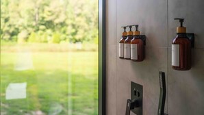 A bathroom with a window view of a green landscape. There are three bottles of liquid soap on a wall-mounted dispenser.