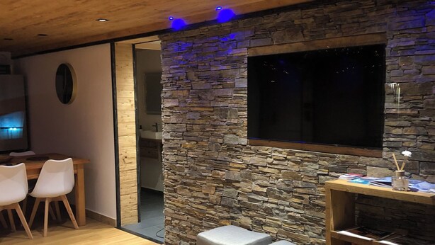 A modern living room with a stone wall, wooden ceiling, and a flat-screen TV mounted on the wall. There are two grey stools and a wooden table with a magazine on it.