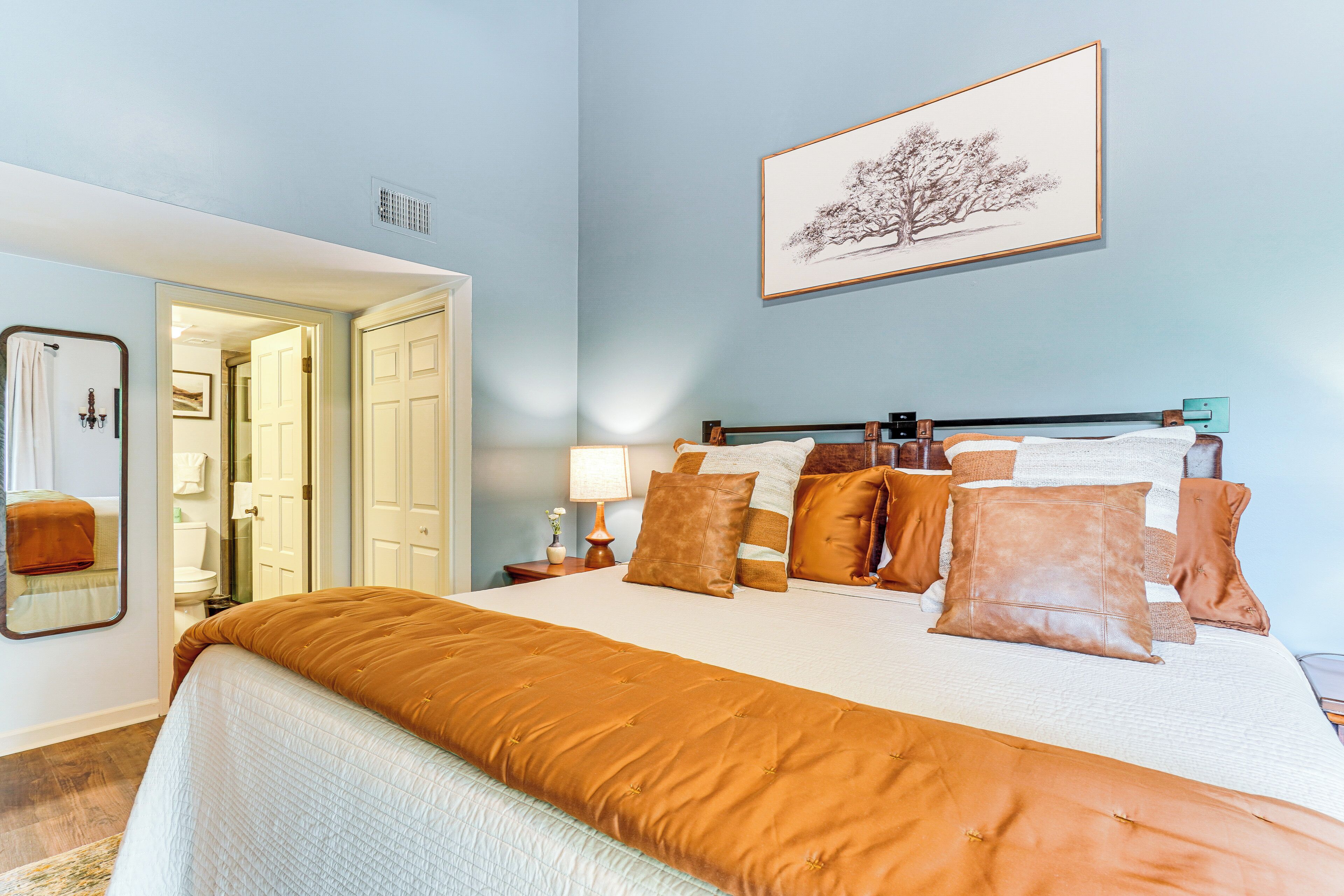 A bedroom with a large bed, a nightstand with a lamp, a framed picture on the wall, and a door leading to another room.