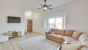 A living room with a brown leather sofa, a flat-screen TV mounted on the wall, a ceiling fan, and a large window providing natural light. - 3 Mi to London Bridge Beach - 'ggs River Retreat' (Lake Havasu City)