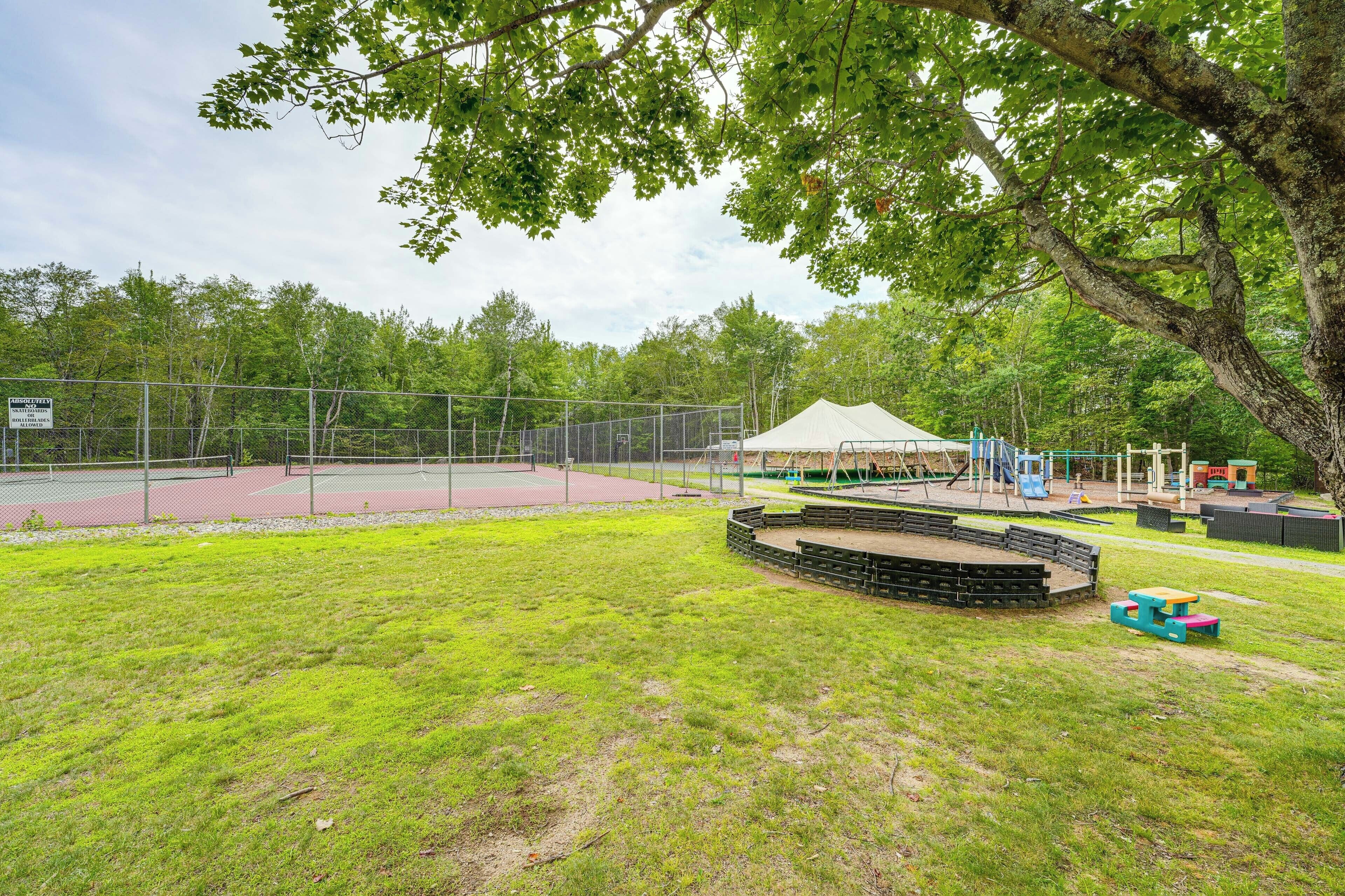Un espacio al aire libre con valla perimetral, que incluye una cancha de tenis, mesas de picnic y un escenario.