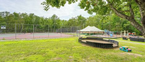 Un espacio al aire libre con valla perimetral, que incluye una cancha de tenis, mesas de picnic y un escenario.