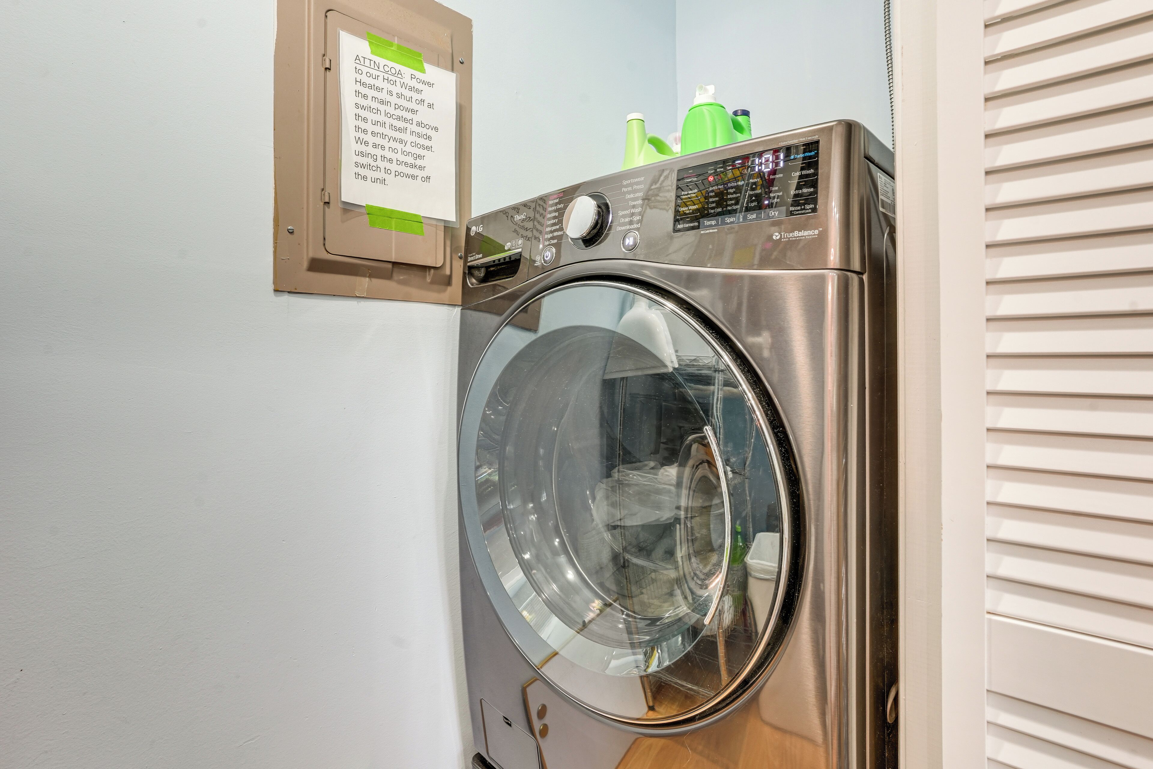 Een was-droogcombinatie met een voorlaadopening, van roestvrij staal, met een bedieningspaneel, een bericht op de muur en een groene fles er bovenop.