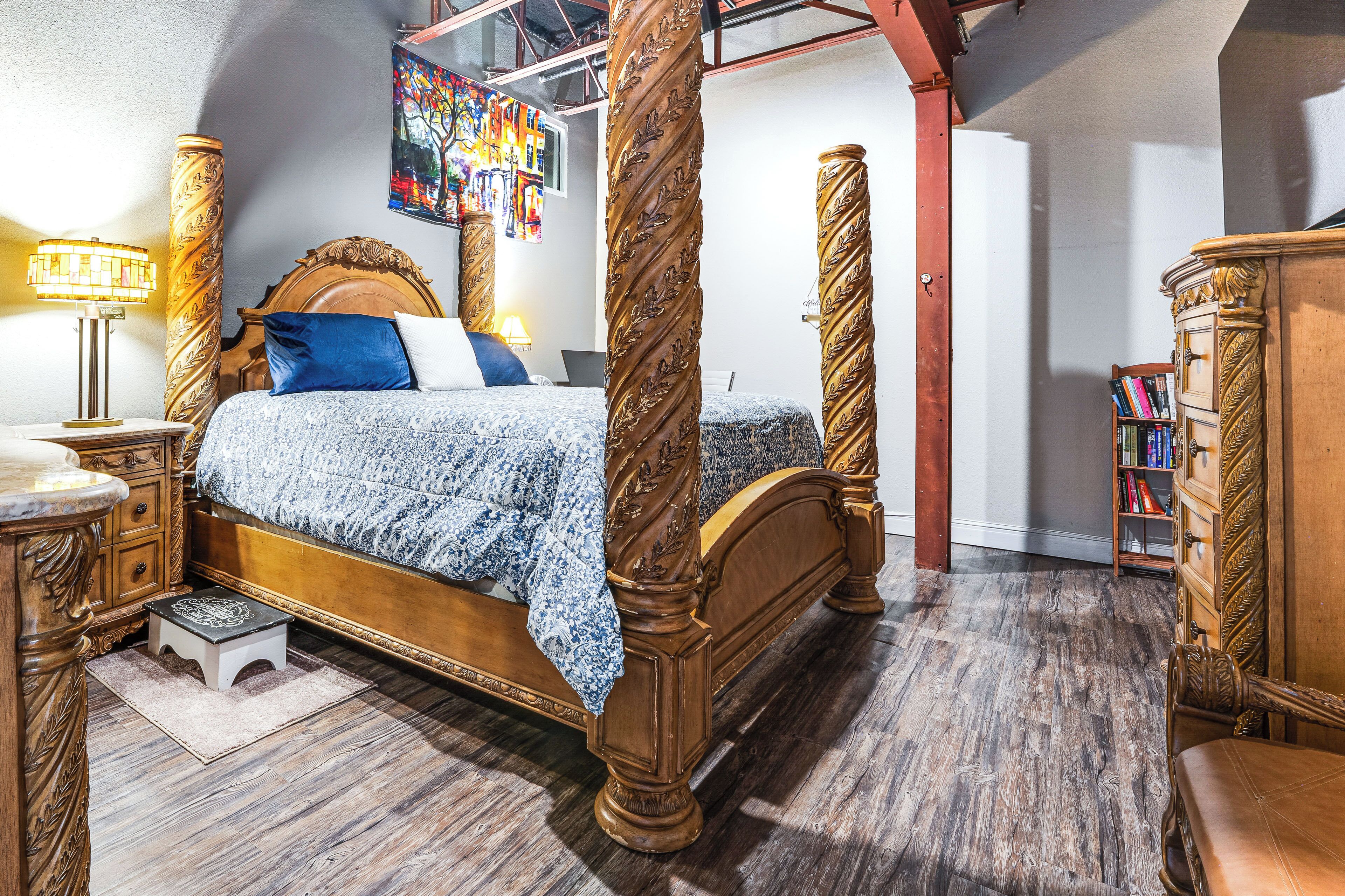 A four-poster bed with a wooden headboard and footboard, a bedside table with a lamp, and a small stool.