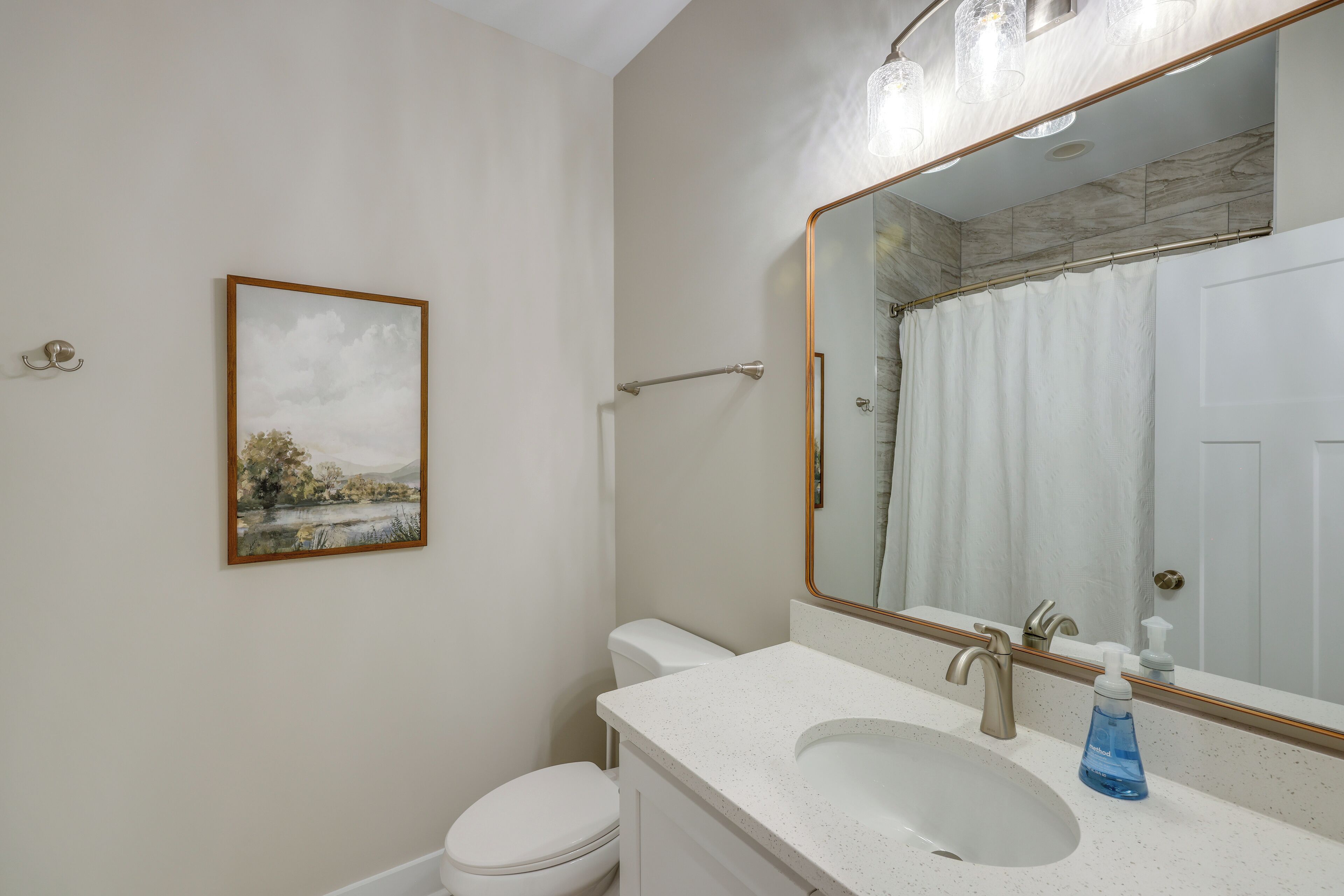 Un bagno con un lavandino bianco, uno specchio grande, un quadro incorniciato con un paesaggio e un water bianco.
