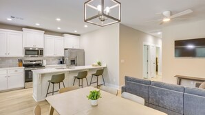Una cocina moderna con armarios blancos, una isla central con taburetes de bar, una mesa de comedor con sillas y un sofá gris en la sala de estar.