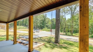 Un porche cubierto con vigas de madera y piso de concreto, con vistas a una zona verde con árboles y un vehículo estacionado.