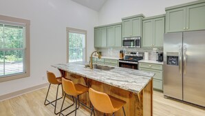 Una cocina moderna con armarios verdes, una isla de mármol con taburetes altos, electrodomésticos de acero inoxidable y ventanas amplias.