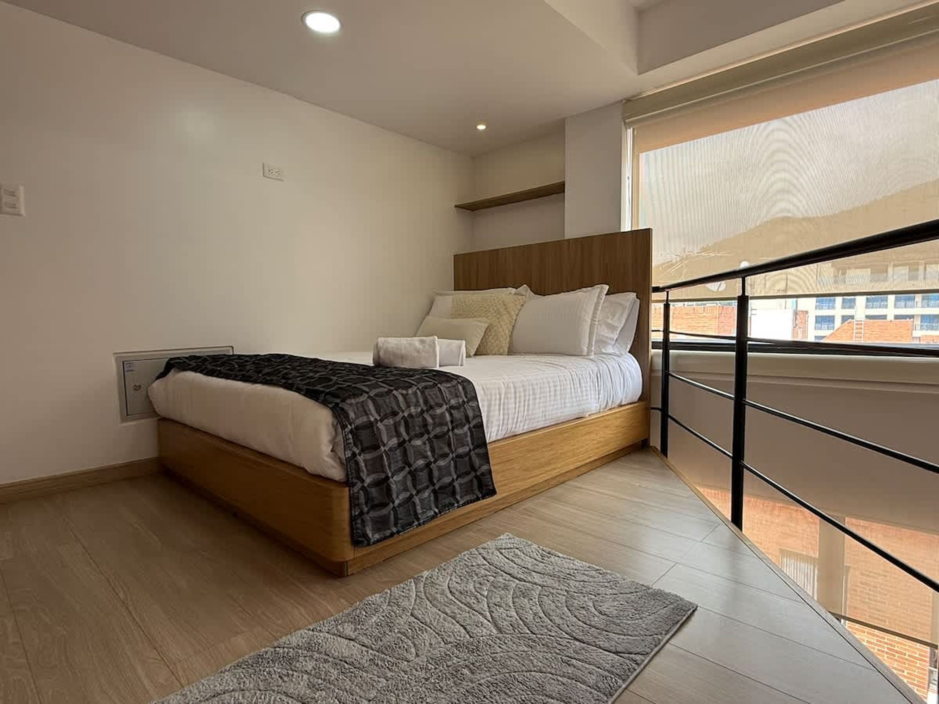 A modern bedroom with a wooden bed, a grey rug, and a view of the city through a balcony railing.