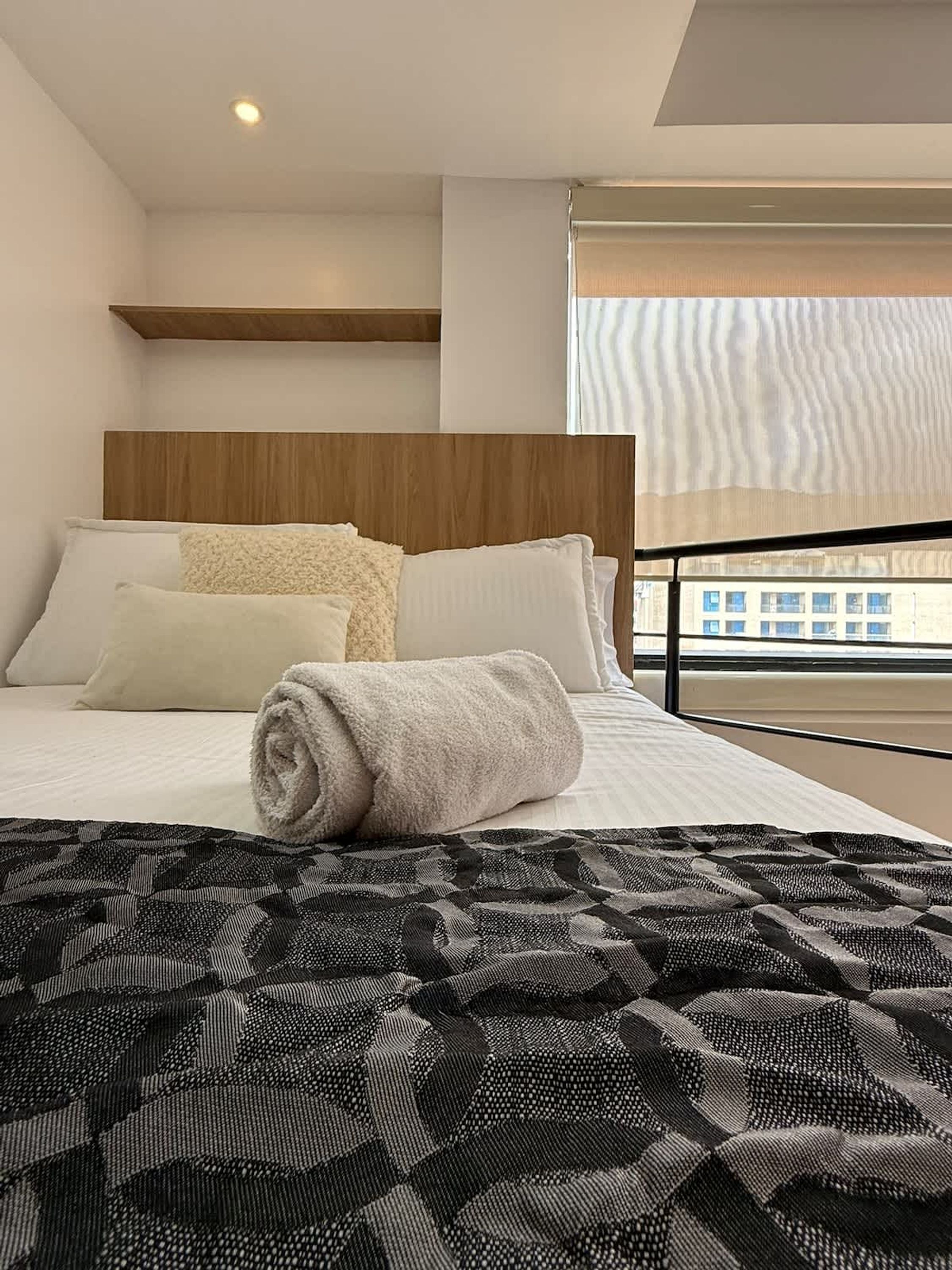 Une chambre d’hôtel avec un lit, une tête de lit en bois, une fenêtre donnant sur des immeubles et une rambarde de balcon.