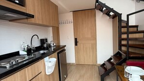 Cocina compacta con microondas, cafetera y pileta. Hay un refrigerador pequeño y una puerta de madera. Una escalera con un cojín rojo conduce a un comedor con una mesa lista para dos.