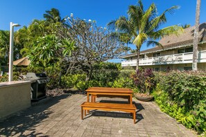 Eine hölzerne Bank in einem Gartenbereich mit Grill und einem Gebäude im Hintergrund.