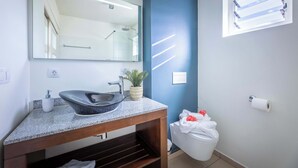 A bathroom with a granite countertop, a vessel sink, a mirror, a towel rack, a toilet, and a window with blinds.