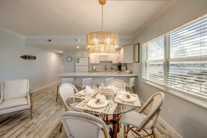 A dining area with a round table set for six, surrounded by wicker chairs, a hanging light fixture, and a view through large windows.