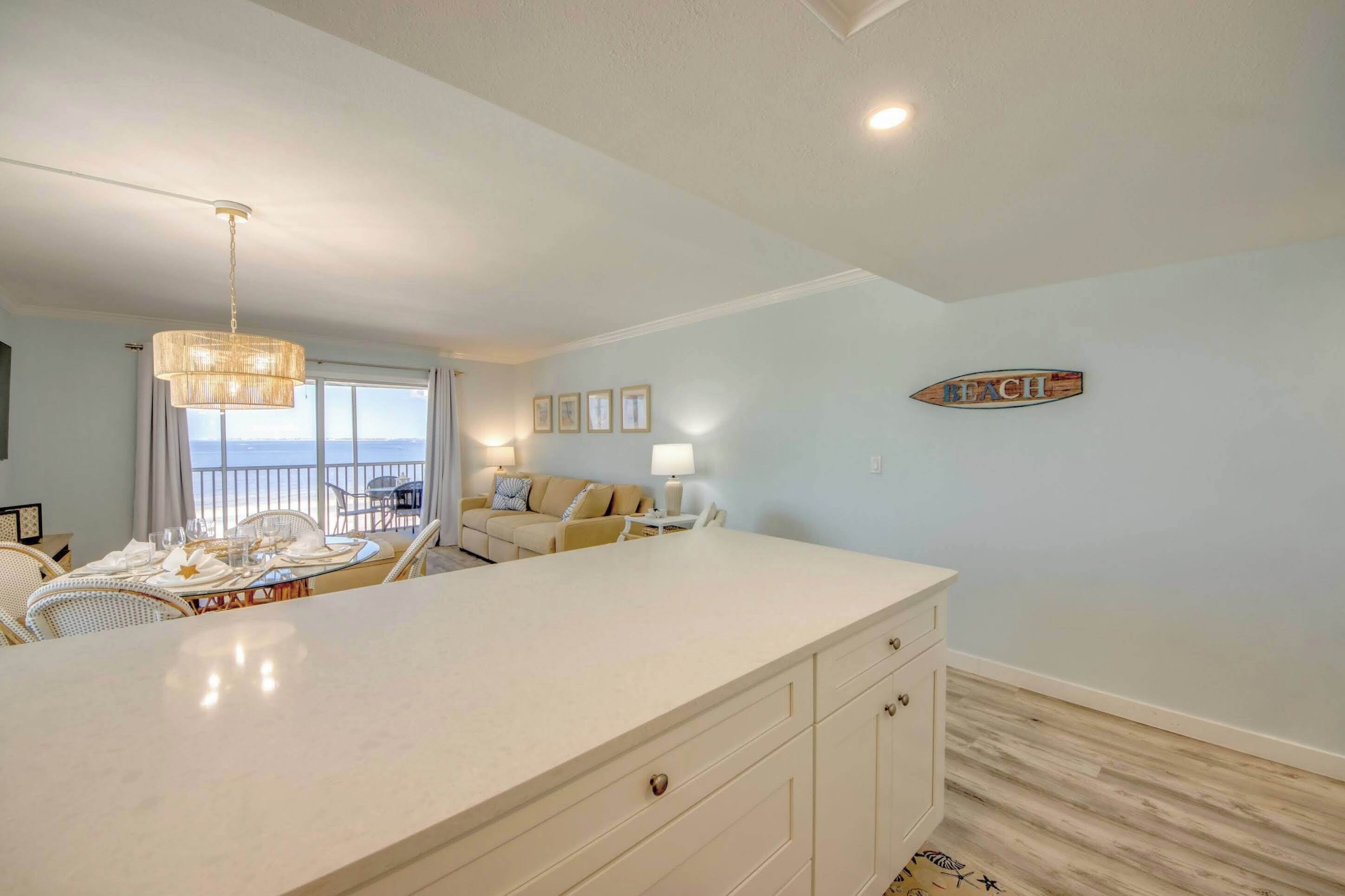 A room with a large white island, a dining table set with white tableware, a sofa, and a view of the ocean.