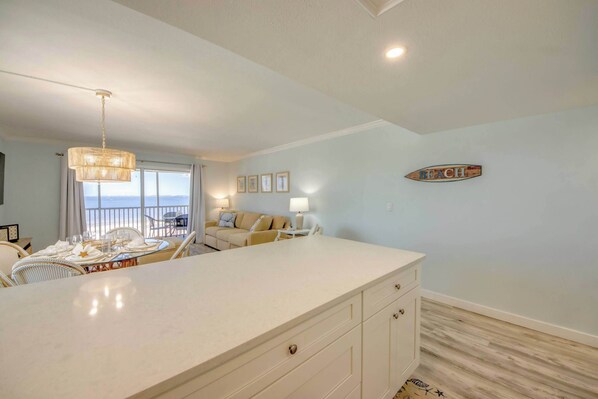 A room with a large white island, a dining table set with white tableware, a sofa, and a view of the ocean.
