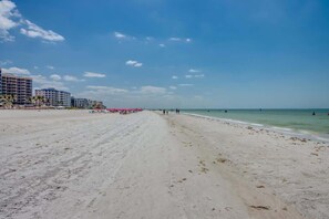 A wide sandy beach with a clear sky, buildings in the background, and a few people in the distance.