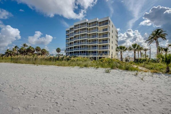 A multi-story hotel with balconies, located near a sandy beach with palm trees.