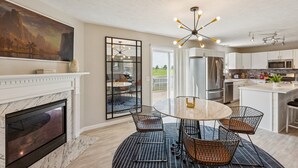 A modern kitchen with a marble fireplace, a dining area with wire chairs, and a large window overlooking a deck.