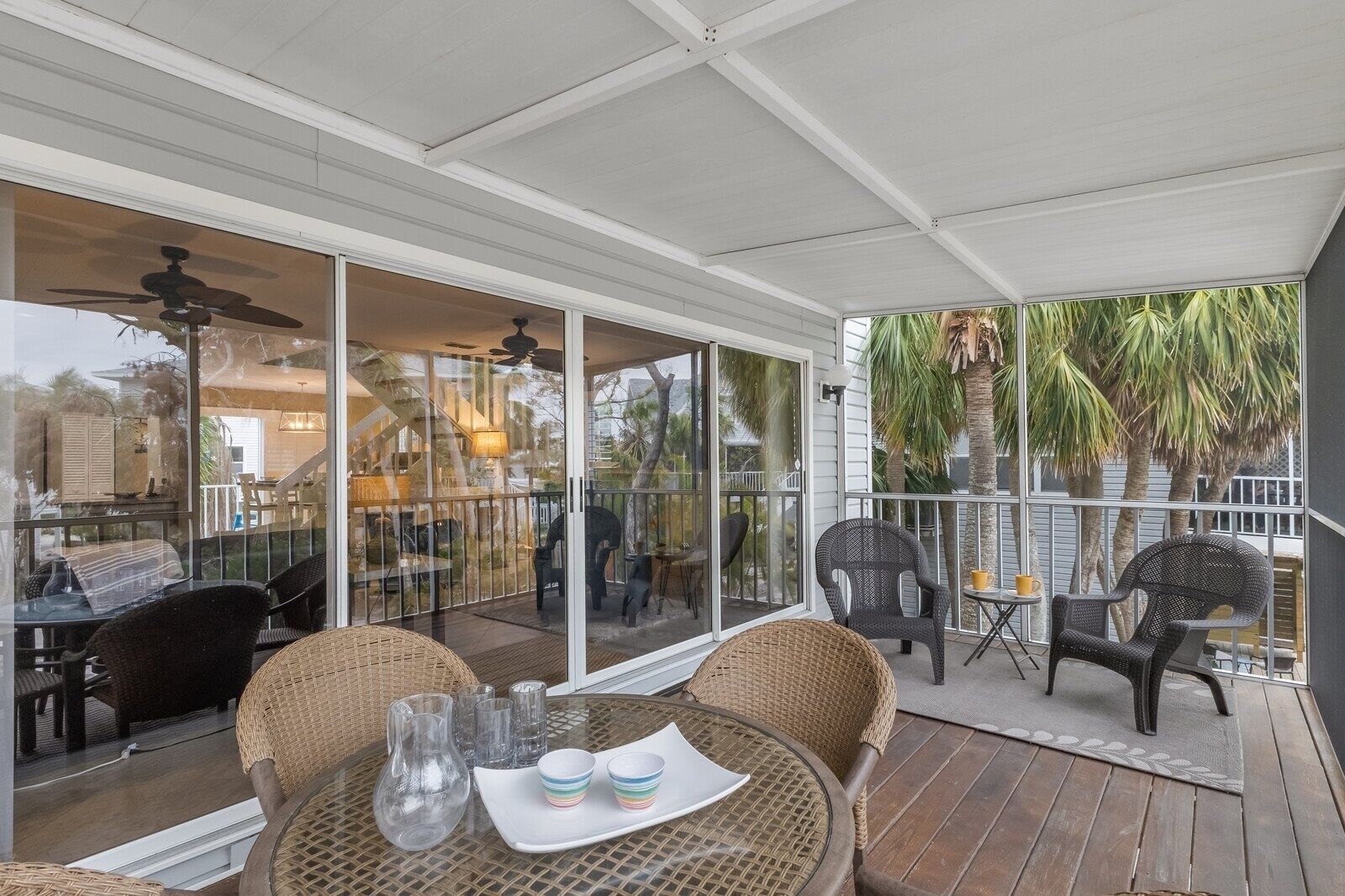 A covered outdoor patio with wicker furniture, glass tables, and a view of a house and palm trees. - North Beach Village #62 3 Bedroom Home (Holmes Beach)