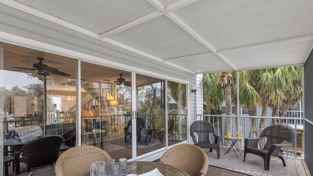 Un patio techado al aire libre con mobiliario de mimbre, mesas de vidrio y vista a una casa y palmeras.