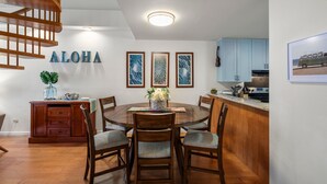 A dining area with a wooden table and chairs, a kitchen with light blue cabinets, and a staircase with wooden railings. - Kamaole Sands #5-401 2 Bedroom Condo (Kihei)