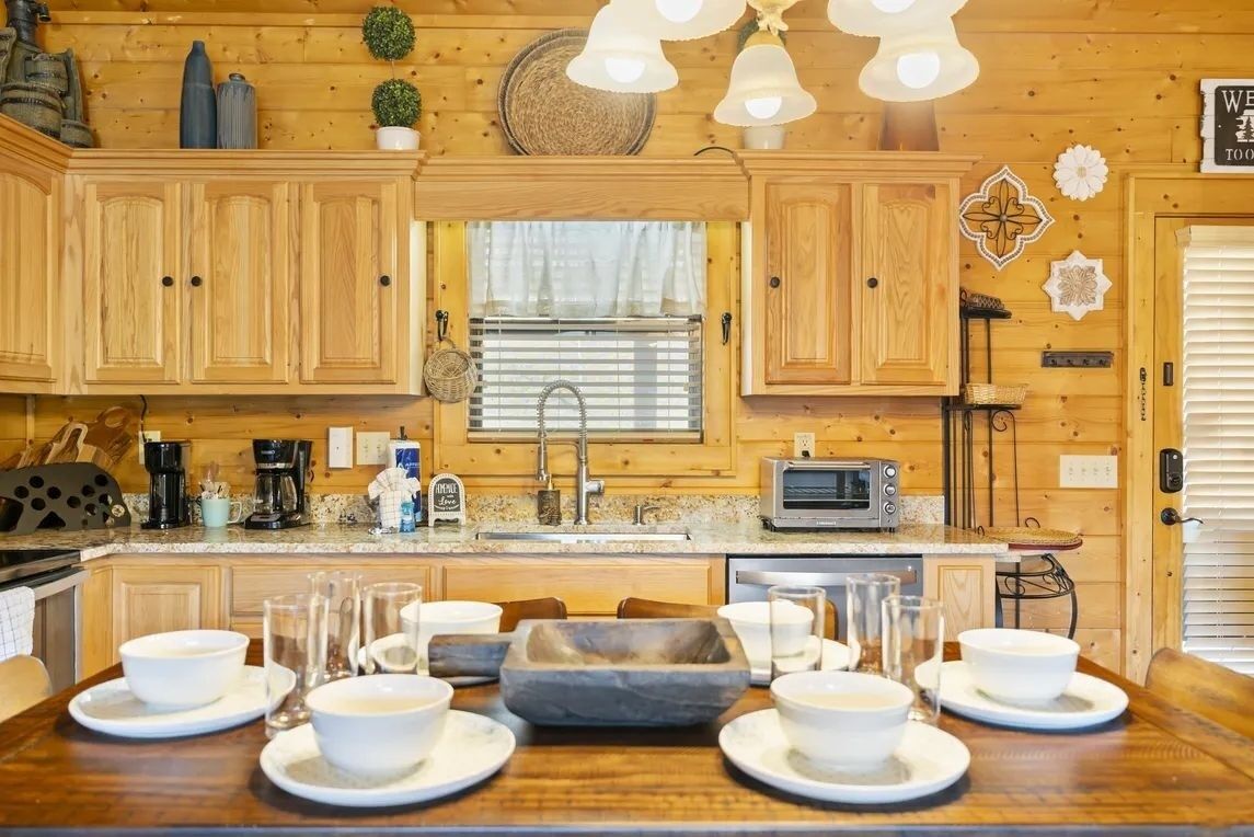 Het interieur van een houten blokhut met een keukenhoek, inclusief houten kasten, een spoelbak, een fornuis en een eettafel gedekt met kommen en glazen.