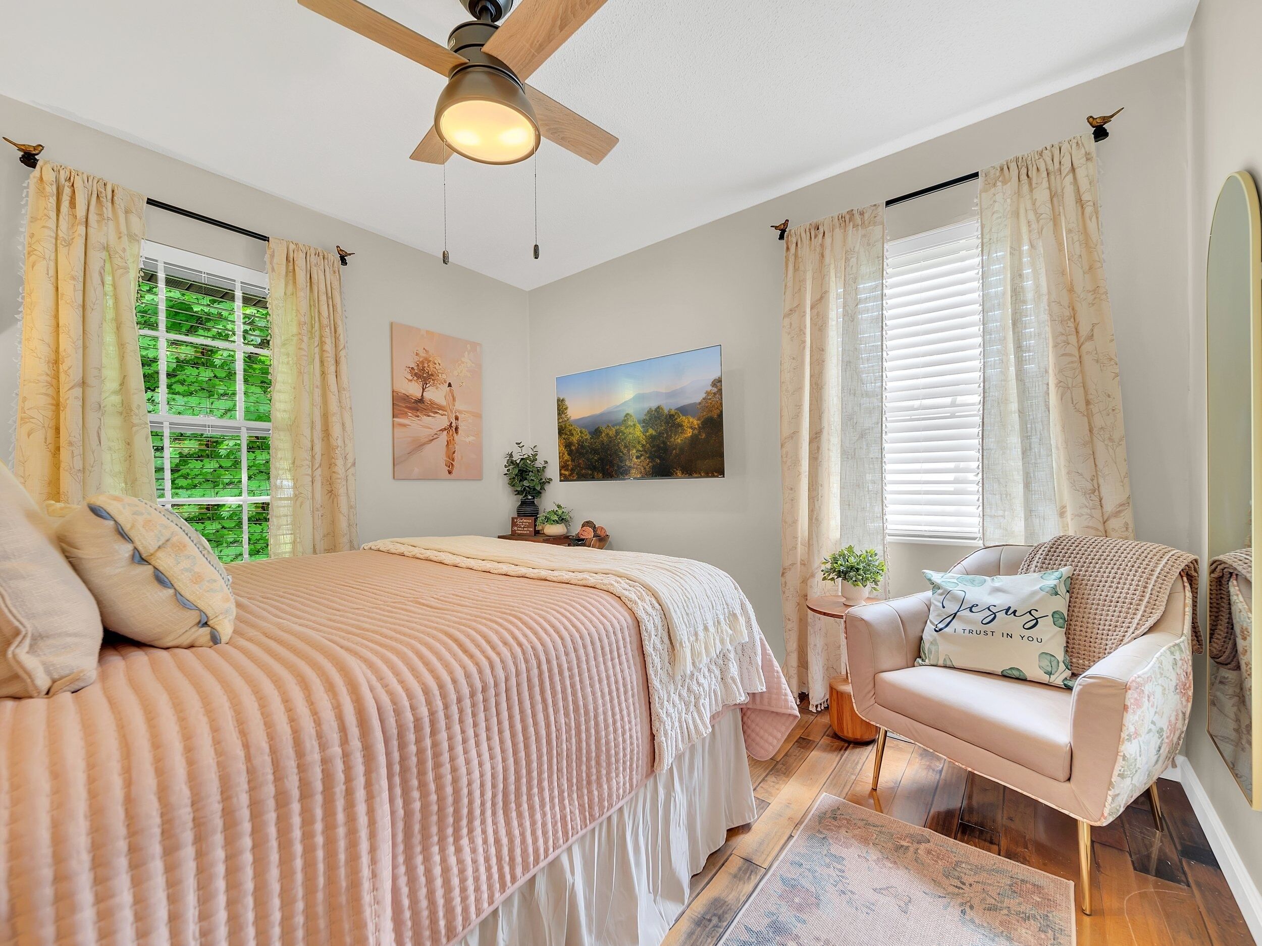 Une chambre à coucher comprenant un lit, une chaise, un ventilateur de plafond, une fenêtre avec des rideaux et un tableau accroché au mur.