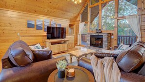 A cozy cabin interior with a fireplace, leather sofas, a flat-screen TV, and large windows. - All Ya Need 4 Bedroom Cabin (Sevierville)