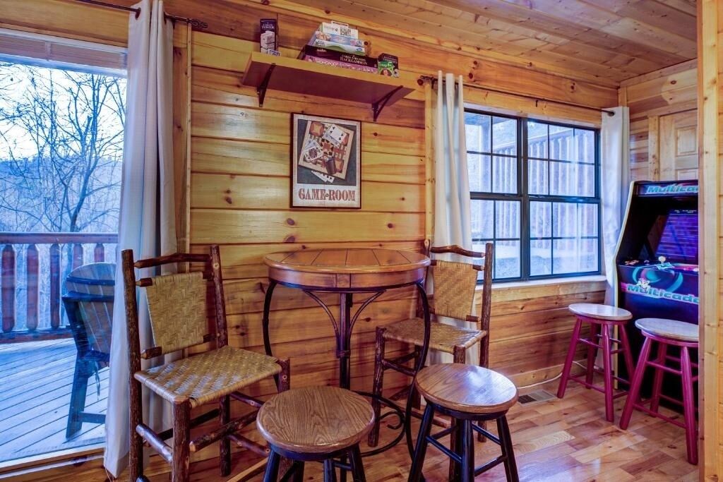 A wooden interior with a 'GAME-ROOM' sign, wooden chairs, a small round table, and a gaming machine.