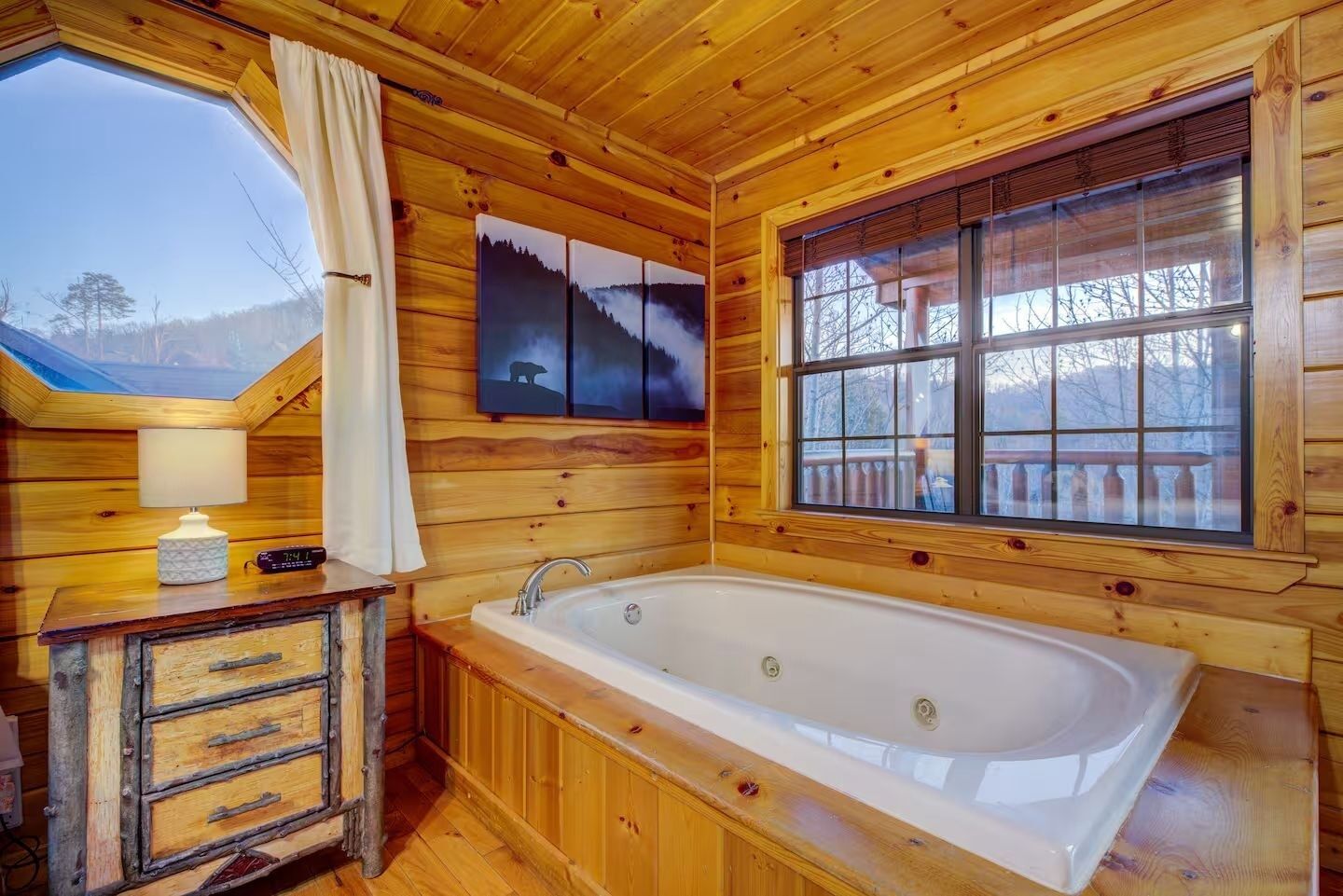 A wooden cabin interior with a large bathtub, a wooden dresser, a window with a view of trees, and a painting on the wall.