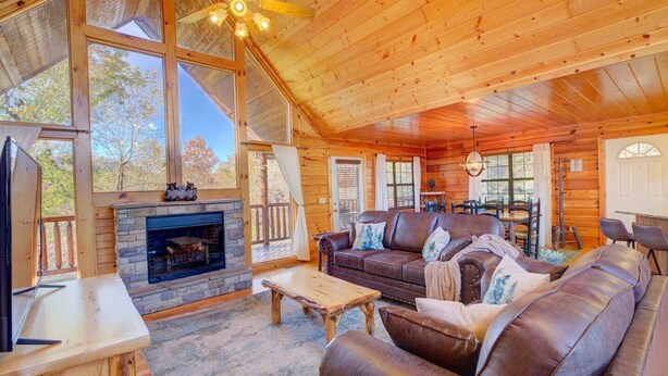 A cozy cabin interior with a stone fireplace, leather sofa, wooden dining table, and large windows offering a view of the outdoors. - All Ya Need 4 Bedroom Cabin (Sevierville)