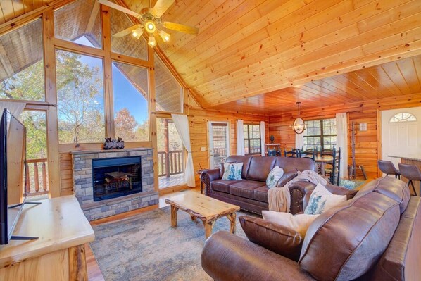 A cozy cabin interior with a stone fireplace, leather sofa, wooden dining table, and large windows offering a view of the outdoors. - All Ya Need 4 Bedroom Cabin (Sevierville)