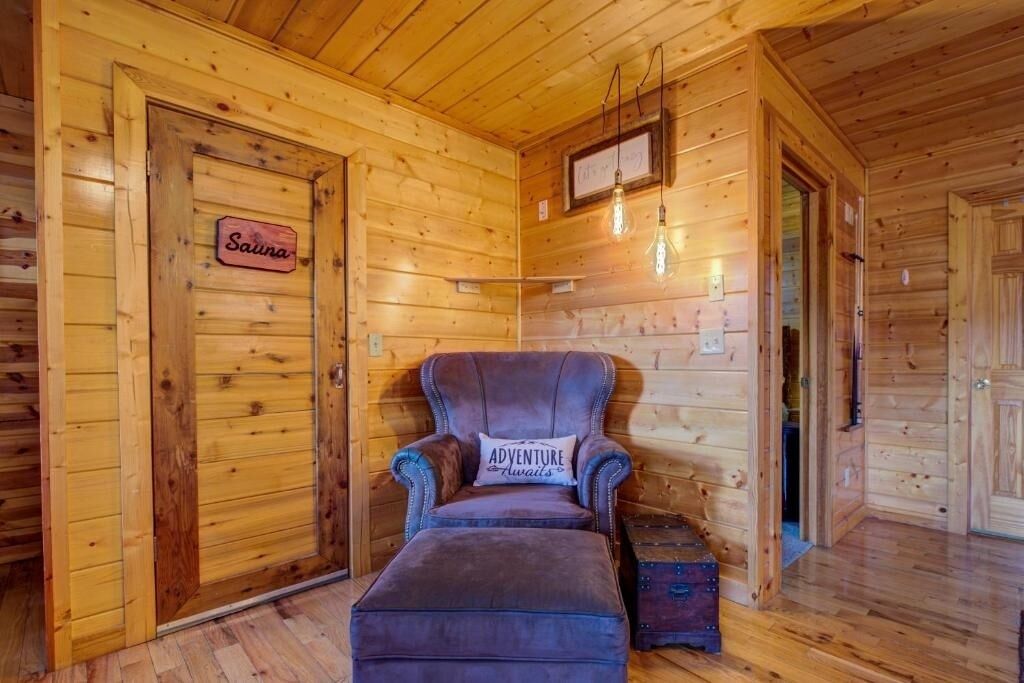 A wooden cabin interior with a sign reading "Sauna" on a door, a plush armchair with a pillow labeled "ADVENTURE Awaits," and a small chest of drawers.